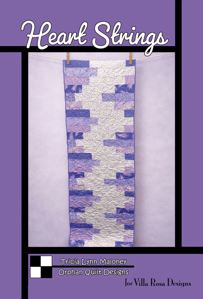 A quilted table runner titled 'Heart Strings' featuring a design of purple and white fabrics, hanging on a clothesline against a light background.