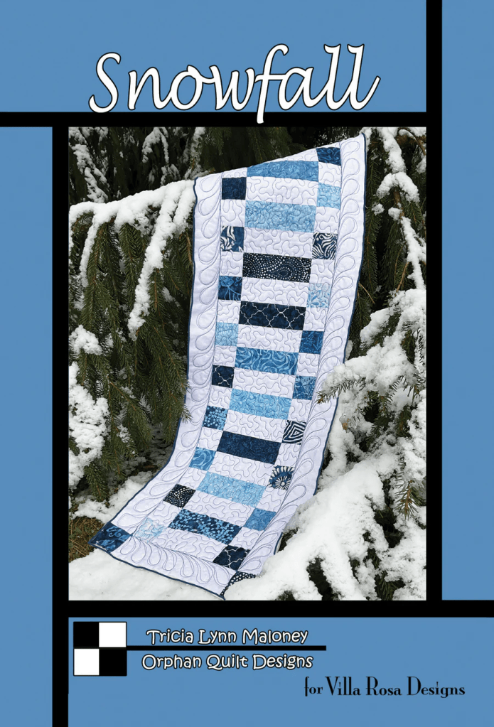 A quilt titled 'Snowfall' designed by Tricia Lynn Maloney, displayed on snow-covered evergreen branches, showcasing a pattern of blue and white fabric pieces.