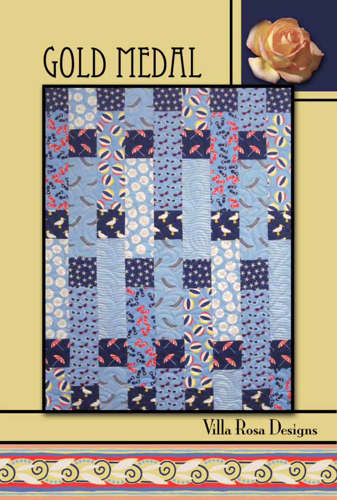Quilt pattern titled 'Gold Medal' featuring a patchwork design in blue tones with various prints, including stars and stripes. The design showcases squares of fabric arranged in a grid, accompanied by a decorative rose in the top corner and branding from 'Villa Rosa Designs' at the bottom.