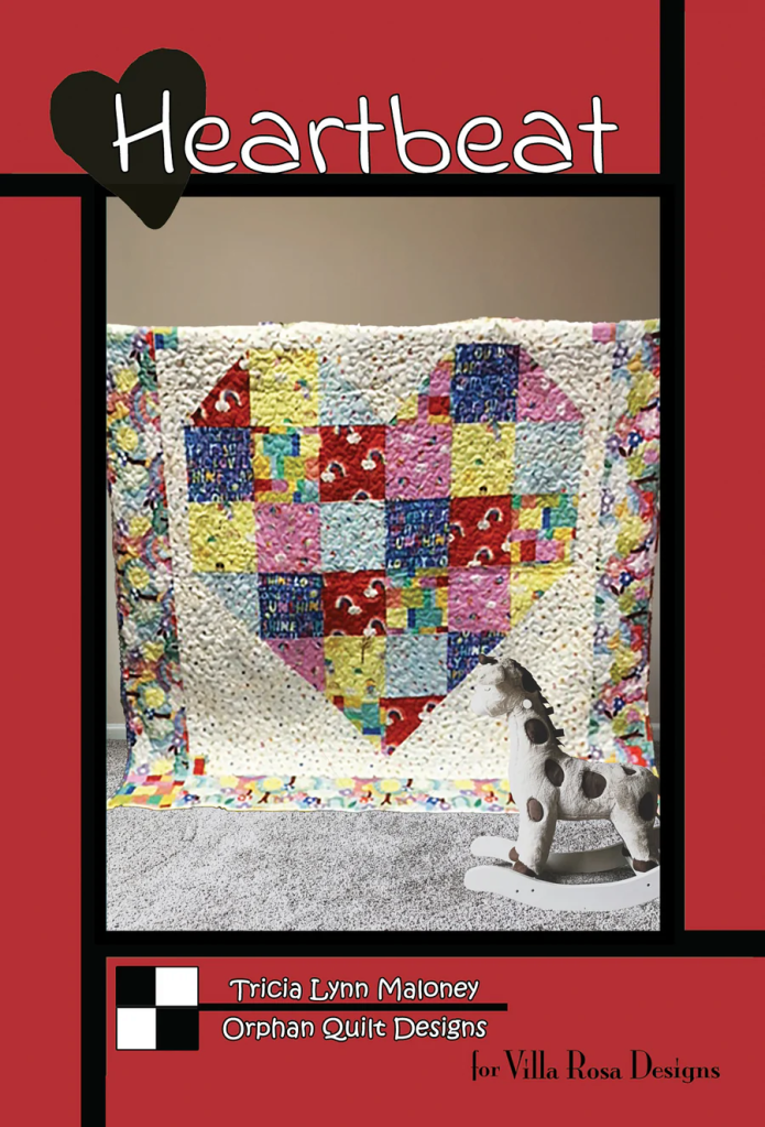 A colorful patchwork quilt featuring a heart design, displayed on a light gray carpet, with a wooden rocking horse toy in the foreground.