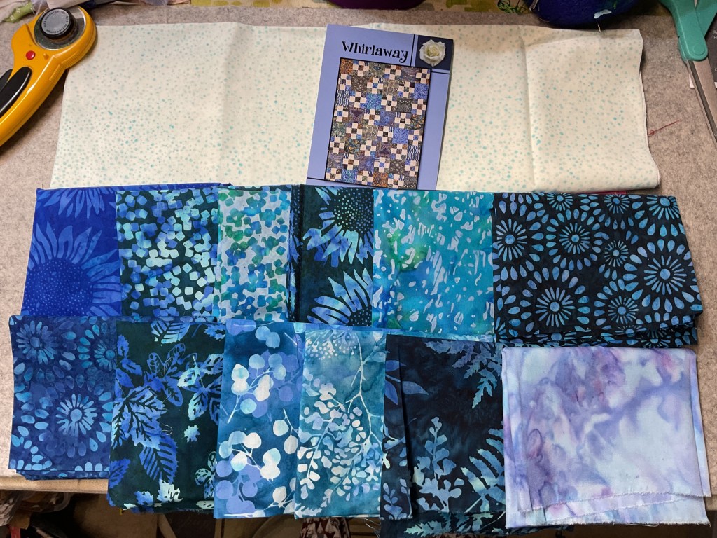 A collection of various blue and green fabric swatches laid out on a table, accompanied by a pattern booklet titled 'Whirlaway'.