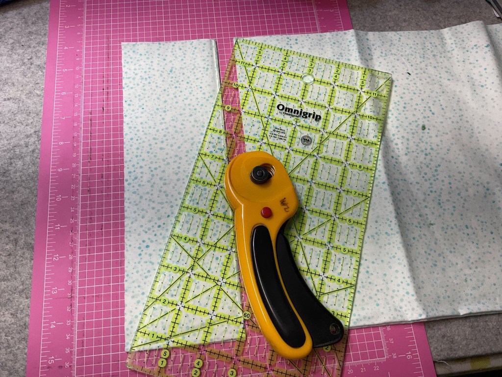 A rotary cutter and a green quilting ruler placed on a pink cutting mat with polka-dotted fabric.
