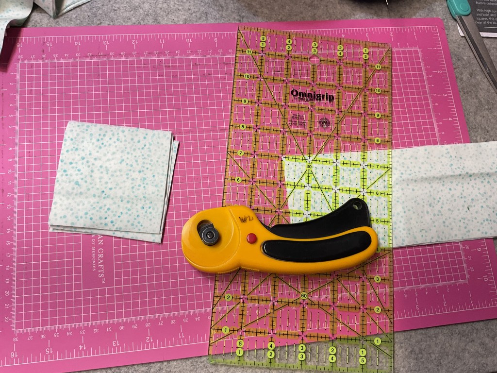 An assortment of fabric squares and a rotary cutter placed on a pink cutting mat with measurement grid lines, indicating a sewing or quilting workspace.
