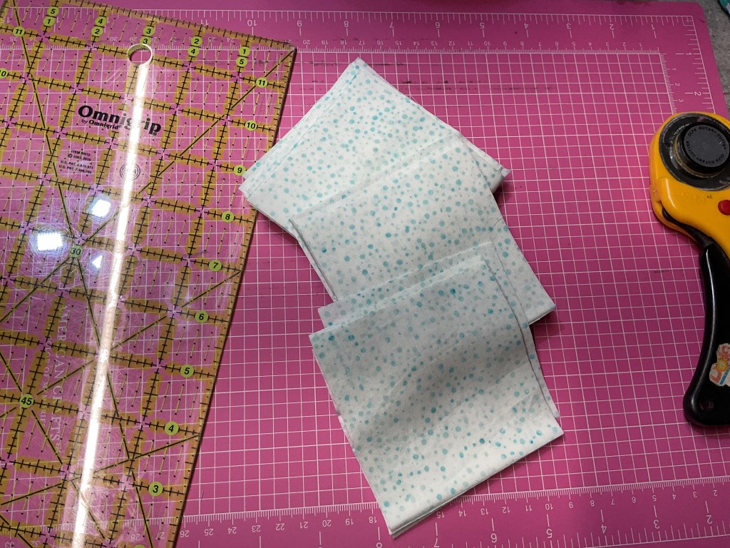 A cutting mat with a transparent ruler and a rotary cutter, alongside several folded pieces of fabric with a light blue polka dot pattern.