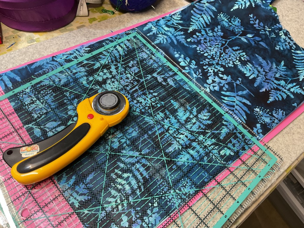 A rotary cutter rests on a cutting mat with green and pink gridlines, beside a piece of fabric featuring blue fern patterns.