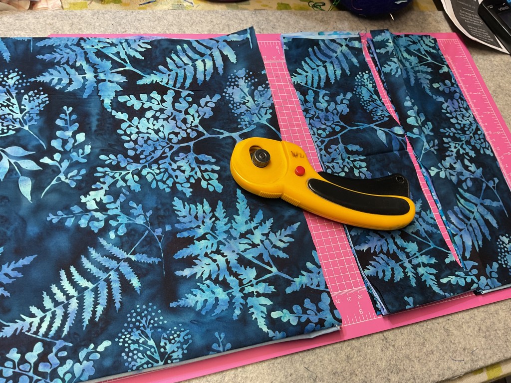 A cutting mat with a vibrant blue and teal fabric featuring a botanical pattern of leaves and ferns. A bright yellow rotary cutter is positioned on the mat next to the fabric.