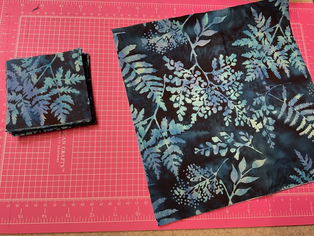 A stack of fabric squares featuring a dark background with blue-green fern and leaf patterns, alongside a larger piece of the same fabric design, laid out on a pink cutting mat.