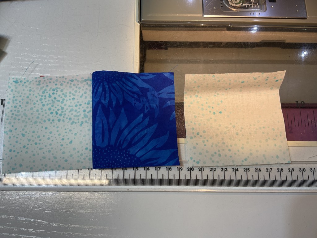 Three pieces of fabric arranged side by side on a sewing table, featuring patterns in light blue, dark blue floral, and a speckled design, with a ruler for measurement.