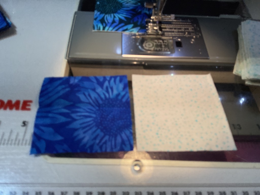 Close-up of two fabric squares placed on a sewing machine surface, one with a blue sunflower pattern and the other with a light speckled design.