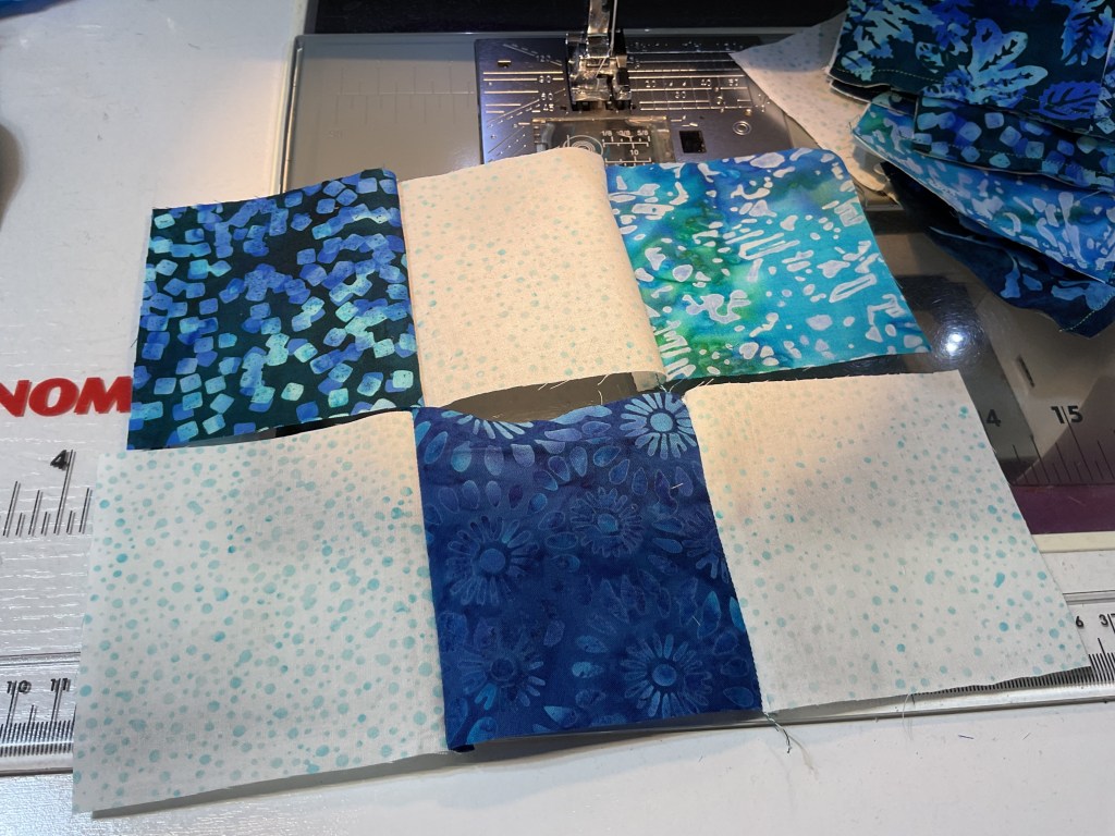 A close-up image of a sewing machine with fabric swatches arranged in a patchwork layout. The swatches feature various shades of blue and green patterns, including floral designs and abstract textures.