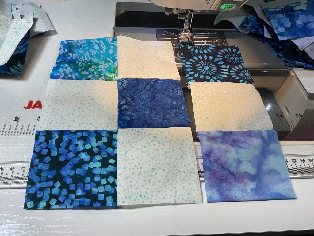 A patchwork of blue and white fabric squares arranged on a sewing machine table, with various patterned fabrics in shades of blue and green.
