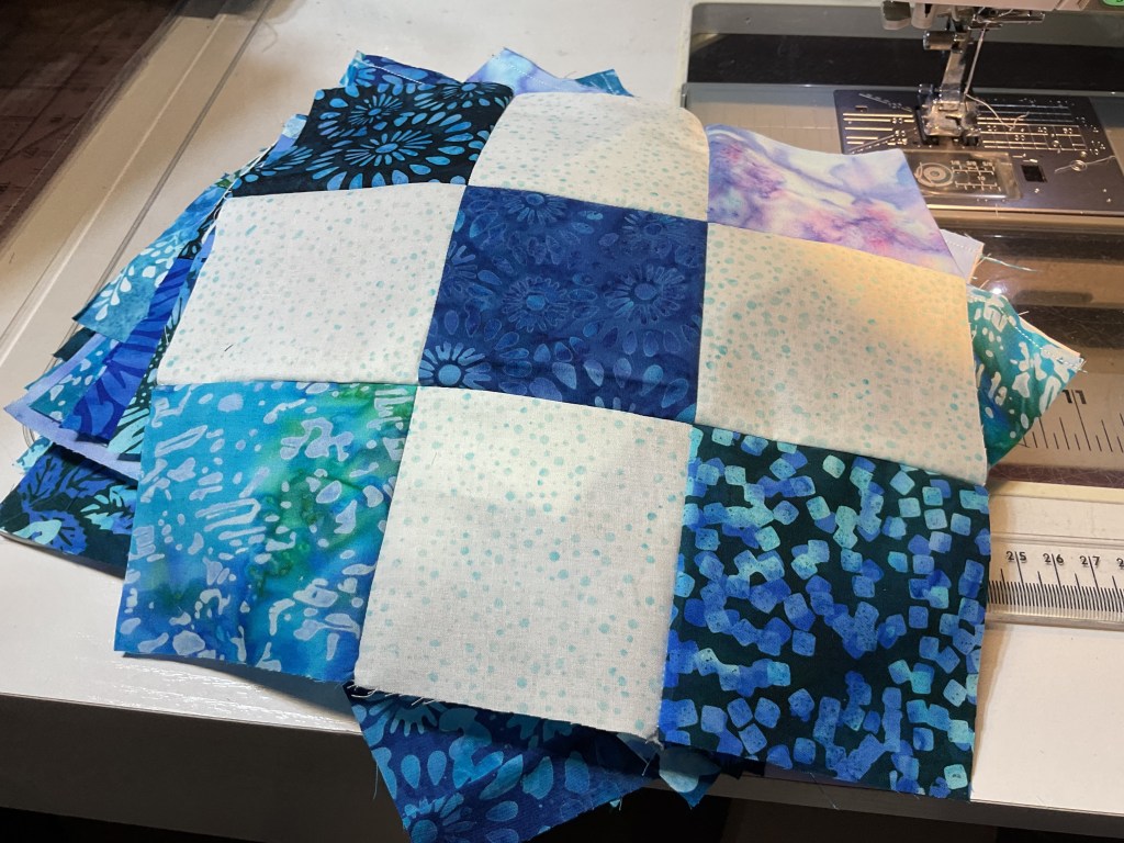 A pile of colorful fabric squares in various shades of blue, arranged in a patchwork design, located next to a sewing machine.
