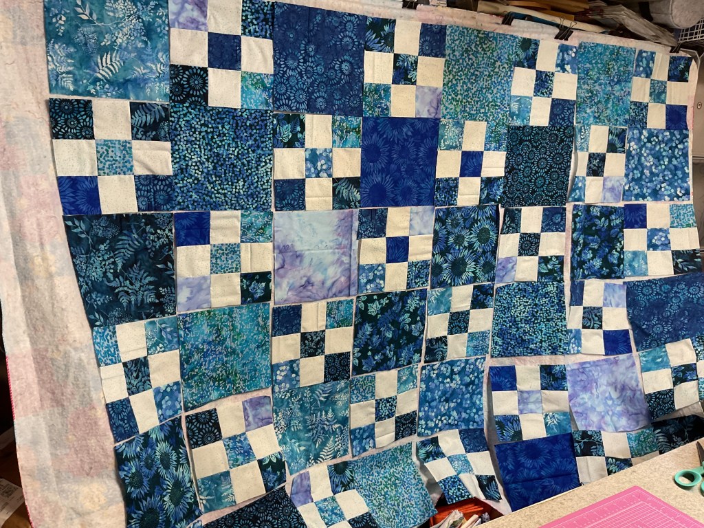 A colorful quilt in progress, featuring a variety of blue and teal fabrics arranged in a patchwork design with white squares.