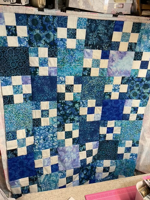 A quilt featuring a patchwork design with various blue and teal fabrics arranged in squares and rectangles, some adorned with floral patterns.