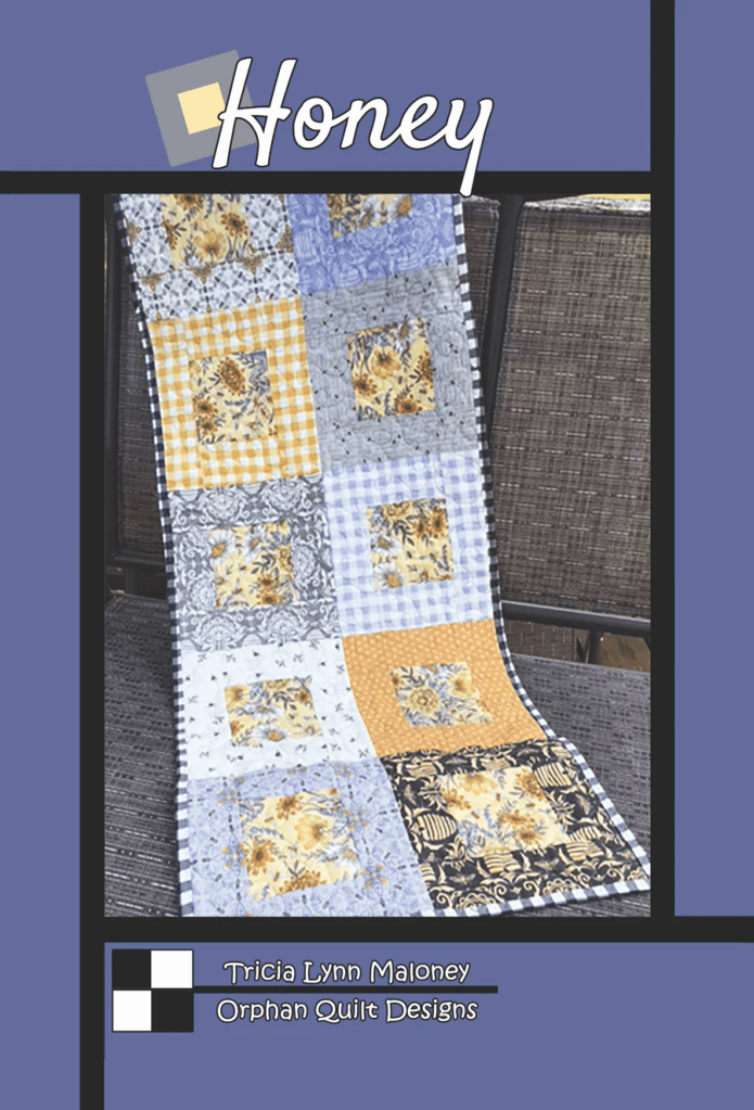 A patchwork quilt with a mix of floral and checkered patterns in yellow, blue, and black, displayed on a dark background with the title 'Honey' and credits to Tricia Lynn Maloney from Orphan Quilt Designs.