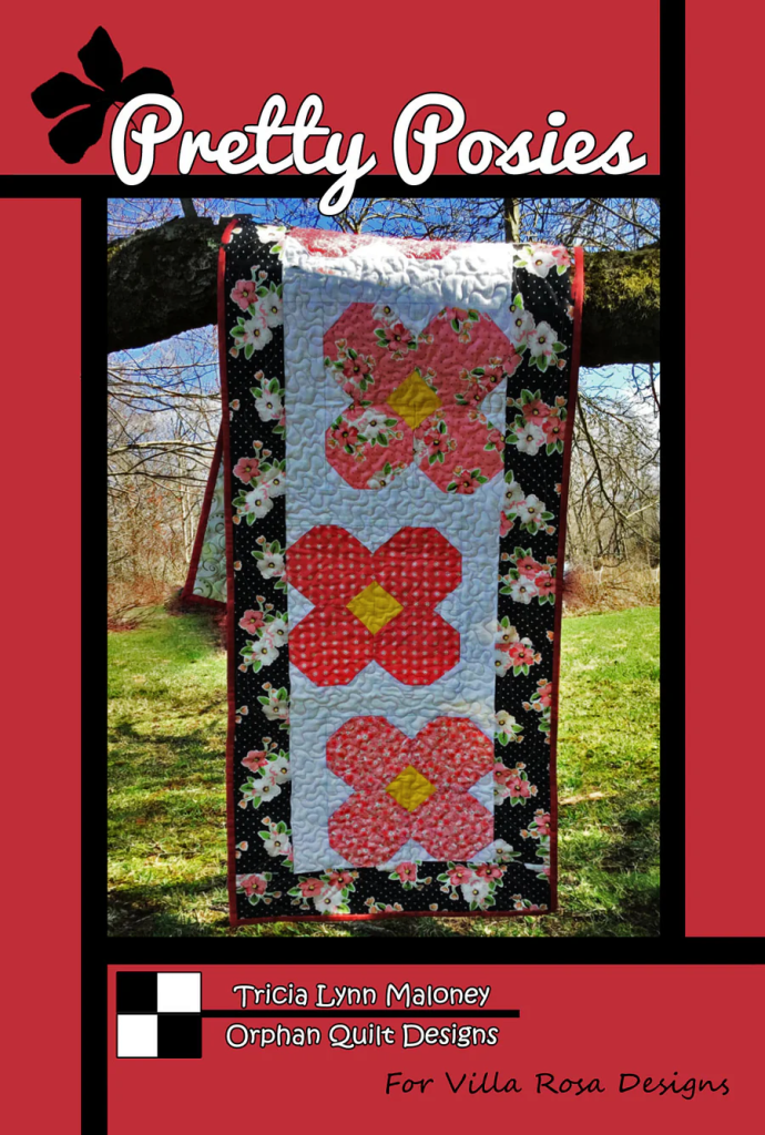 A quilt titled 'Pretty Posies' featuring large colorful floral patterns, hanging on a tree branch in a natural setting. The quilt design incorporates pink, red, and yellow flowers, with a black and white floral border.