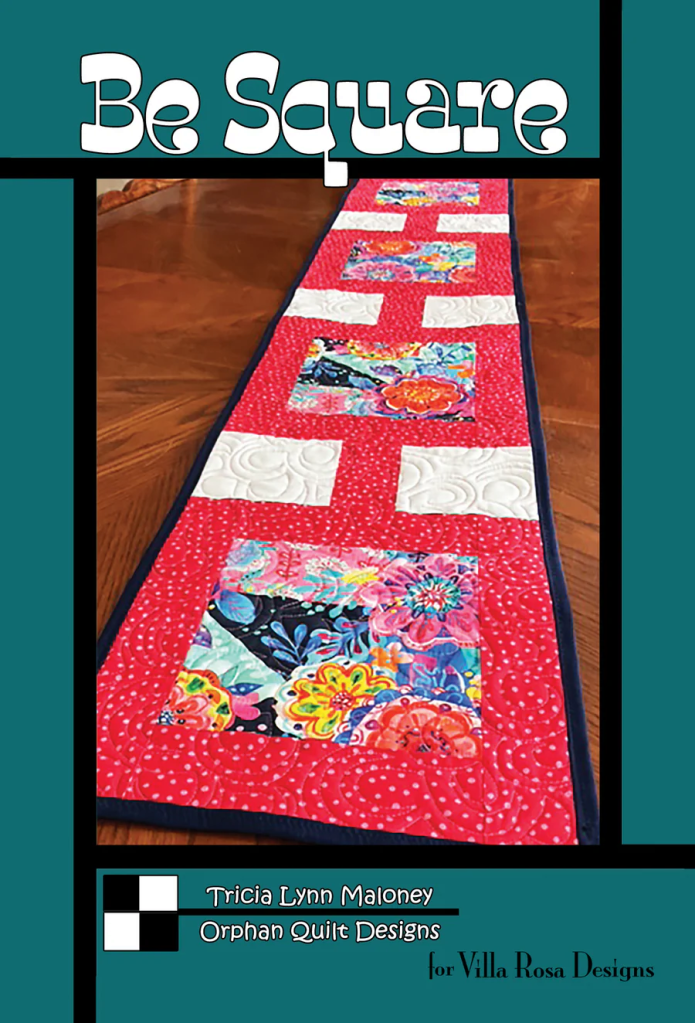 A colorful quilted table runner featuring square blocks with floral patterns on a red background, titled 'Be Square' by Tricia Lynn Maloney. The design includes white spaces between the colorful squares.