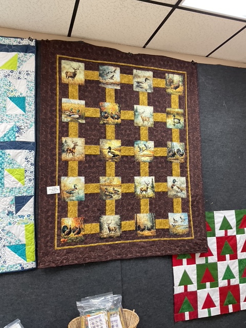 A wall hanging quilt featuring various nature-themed fabric squares including images of animals and birds, bordered with a dark brown fabric and yellow highlights.
