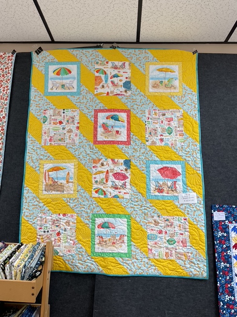 Colorful quilt featuring beach-themed blocks with umbrellas, beach chairs, and seaside illustrations, arranged in a diagonal pattern against a yellow and light blue background.