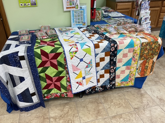 A display of colorful quilts on a table, showcasing various patterns and fabrics, with sewing supplies and patterns arranged nearby.