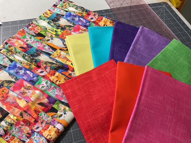 An assortment of colorful fabric pieces laid out on a cutting mat, featuring a vibrant patterned fabric with cat illustrations in the background.