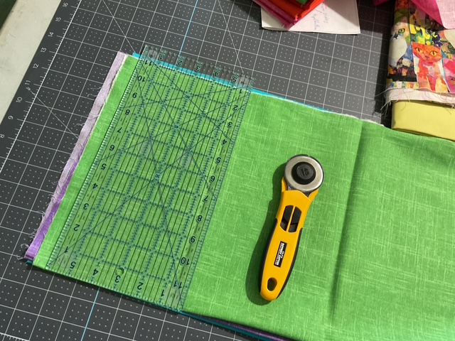 A cutting mat featuring a green fabric square, a transparent ruler, and a rotary cutter, arranged for quilting or sewing projects.