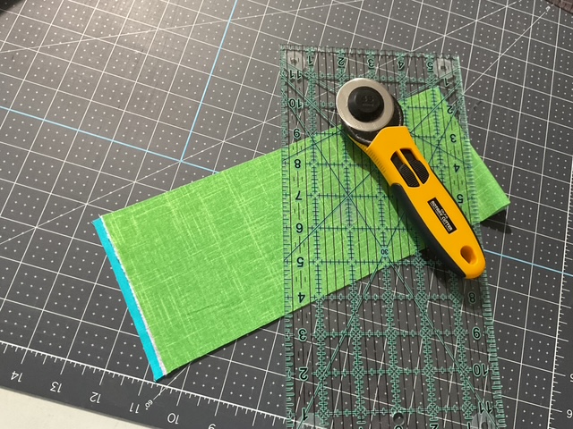 A rotary cutter and clear ruler are placed on a cutting mat with a piece of green fabric laid out for cutting.