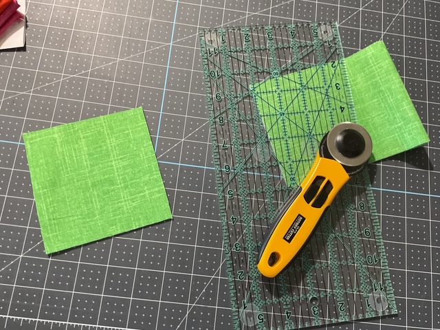 A rotary cutter lies on a cutting mat with two green squares of fabric, one partially cut.