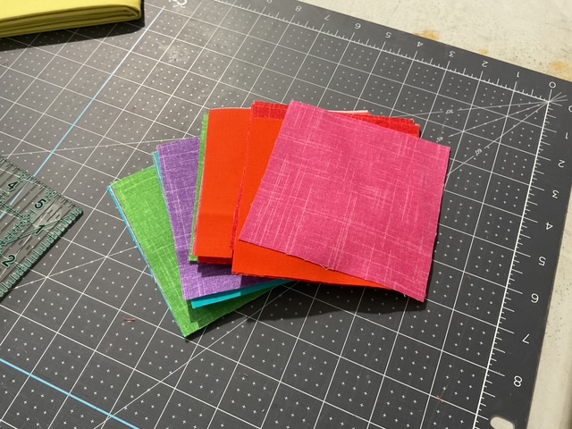 A stack of colorful cloth squares in various shades including pink, red, green, and blue, arranged on a cutting mat with a grid pattern.