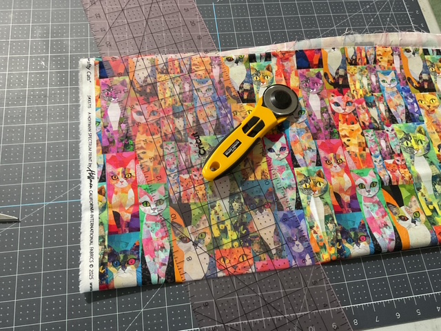 A colorful fabric featuring cat designs laid on a cutting mat, accompanied by a rotary cutter and a clear ruler for measuring.