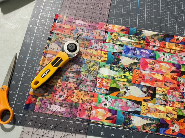 A rotary cutter and scissors on a cutting mat with colorful cat-themed fabric being cut with a ruler.