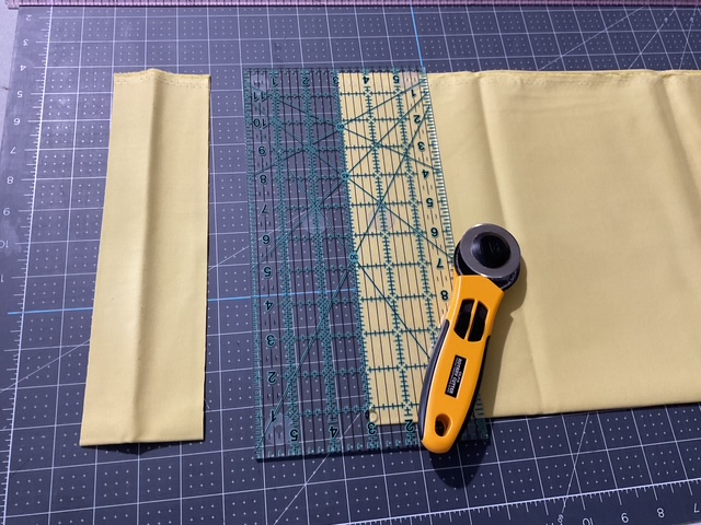 Two pieces of yellow fabric with a transparent ruler and a rotary cutter on a cutting mat.