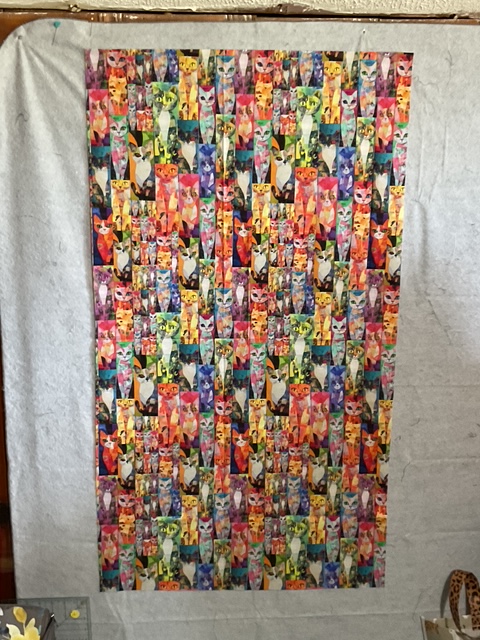 A vibrant and colorful fabric featuring a repeating pattern of stylized cat faces in various colors and expressions.