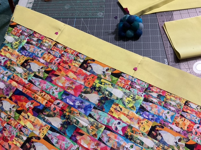 A colorful fabric with a pattern of cats in various artistic styles, bordered by a yellow fabric strip. Pins are used to secure the pieces on a cutting mat.