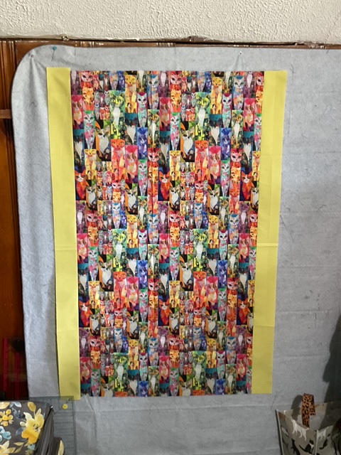 A colorful fabric panel featuring a repeating pattern of cats in various colors and styles, bordered with a yellow edge.