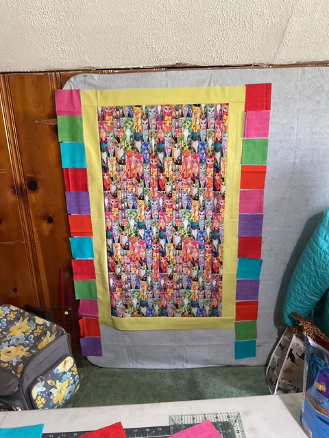 A colorful quilt hanging on a wall, featuring a central panel with vibrant, patterned fabric surrounded by a bright yellow border and accented with squares of various colors along the sides.