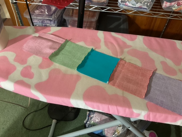 An ironing board covered in a pink cow print fabric, with several fabric swatches in pastel colors including pink, green, blue, and purple laid out on top.