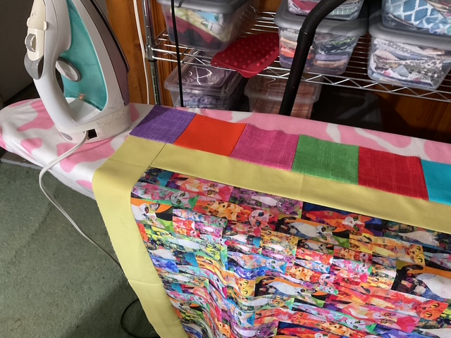 An ironing board with a colorful fabric patchwork spread across it, next to a steam iron. The fabric features various patterns in vibrant colors, and the ironing board cover has a pink and white design.