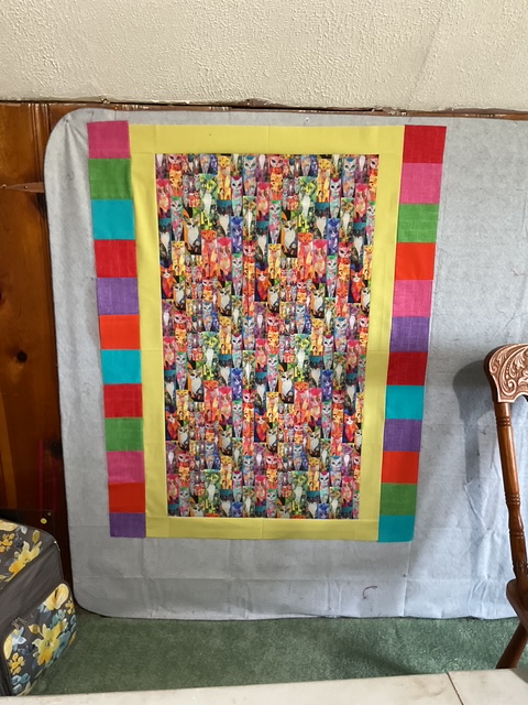 A colorful quilt hanging on a wall, featuring a vibrant pattern of mixed floral and artistic designs surrounded by a bright yellow border and pieced colorful stripes.