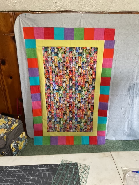 A colorful quilt or fabric panel displayed against a neutral backdrop, featuring a vibrant inner pattern surrounded by a multicolored border.