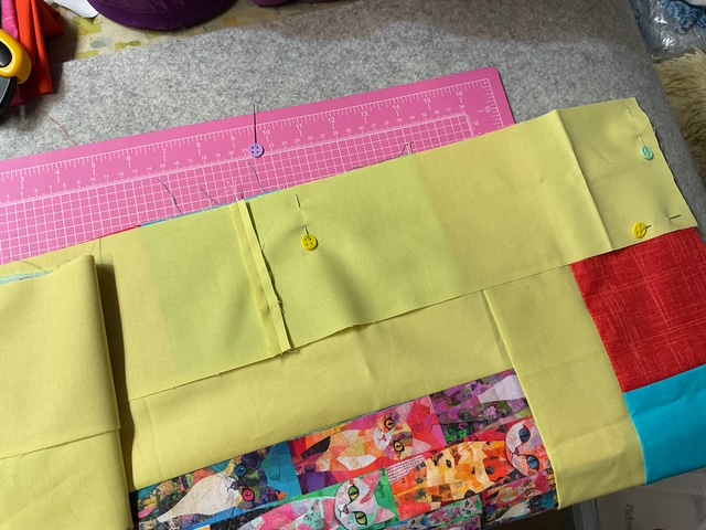 A flat lay of a quilting project featuring a yellow fabric piece being pinned to colorful patchwork fabric on a cutting mat.