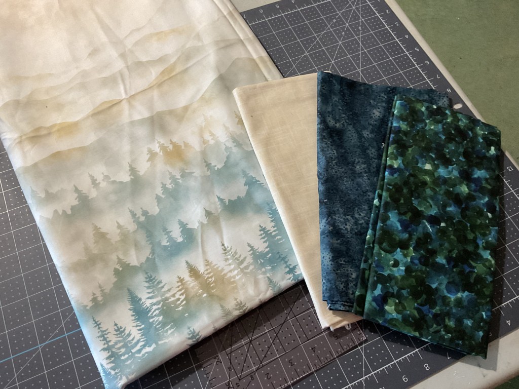 Various fabric pieces displayed on a cutting mat, featuring a scenic landscape design with trees, alongside solid and patterned fabrics in shades of blue and green.
