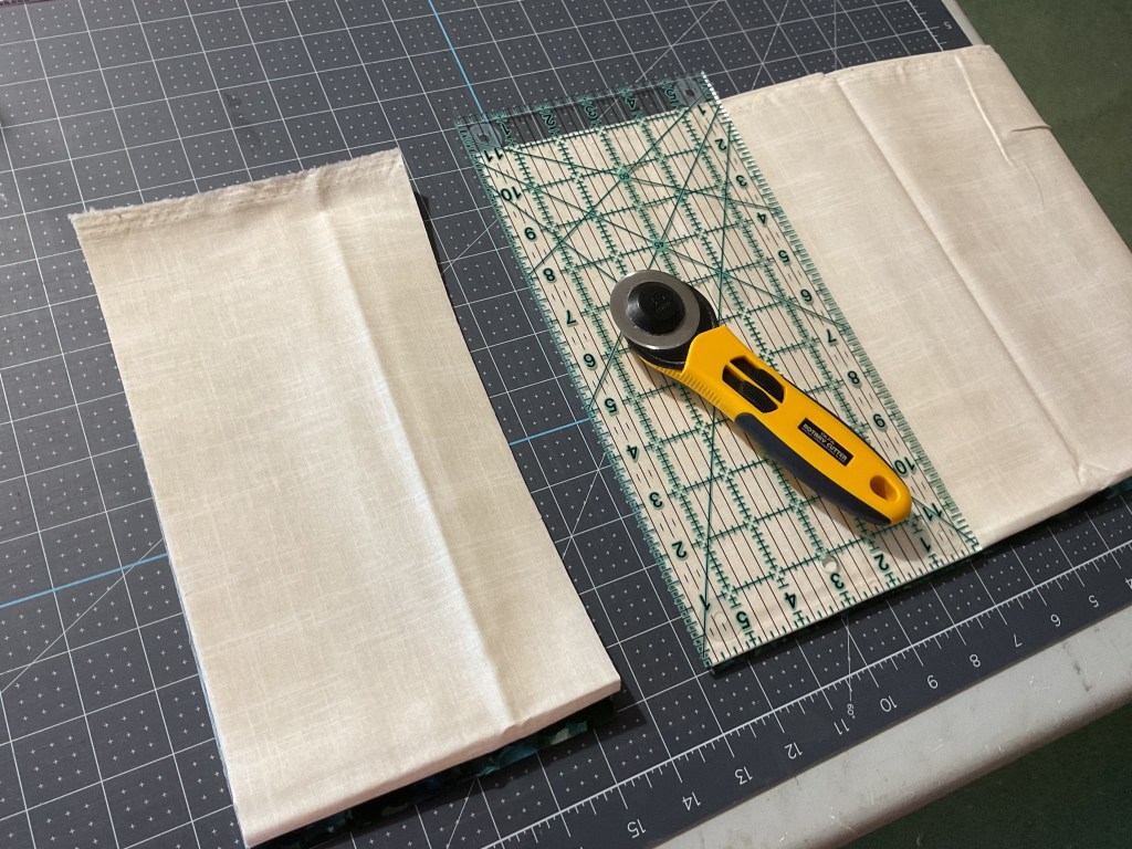 A rotary cutter placed on a cutting mat next to pieces of beige fabric and a measuring ruler marked with grid lines.
