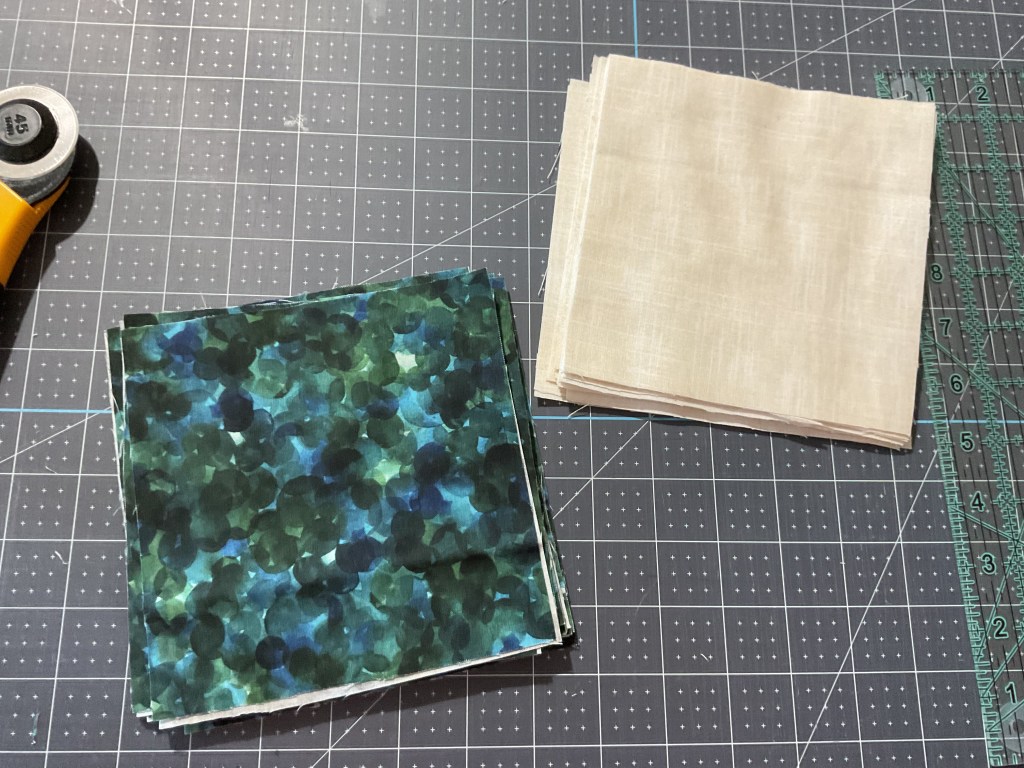 A stack of colorful fabric squares in shades of green next to a stack of beige fabric squares, placed on a cutting mat with a rotary cutter and ruler.