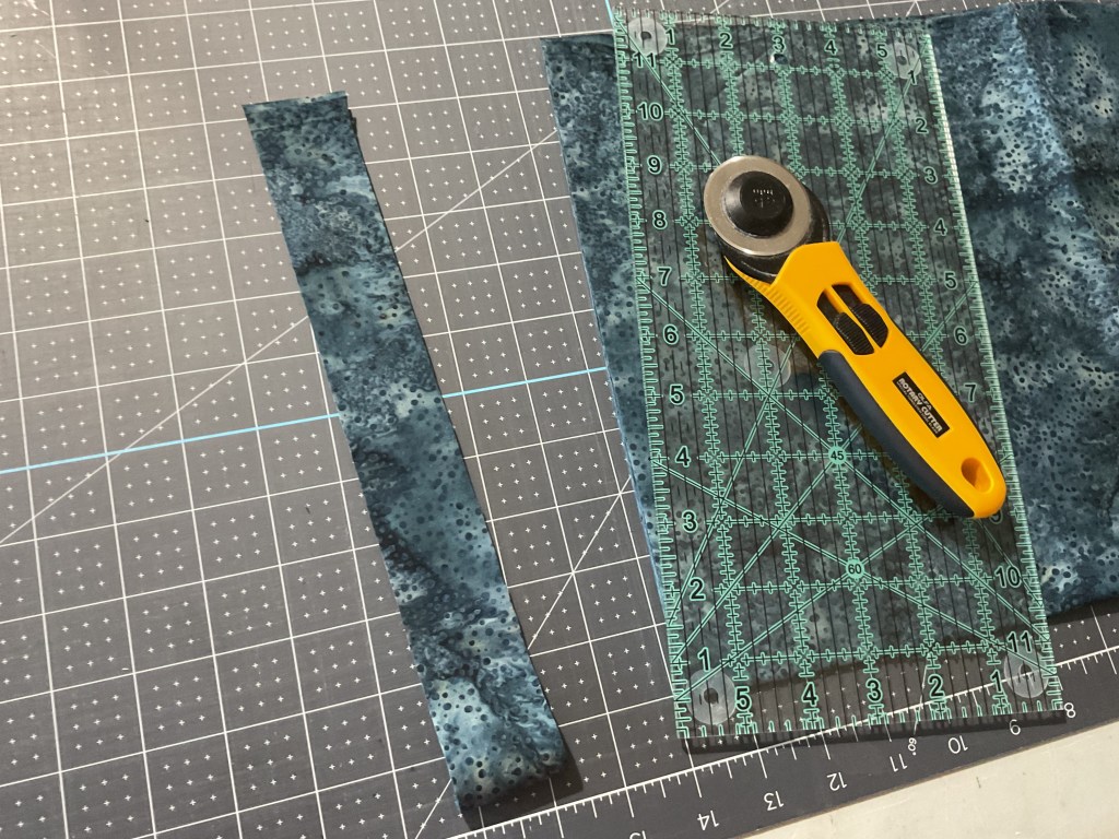 A rotary cutter and a clear ruler on a cutting mat with blue and dark patterned fabric pieces, including a cut strip.