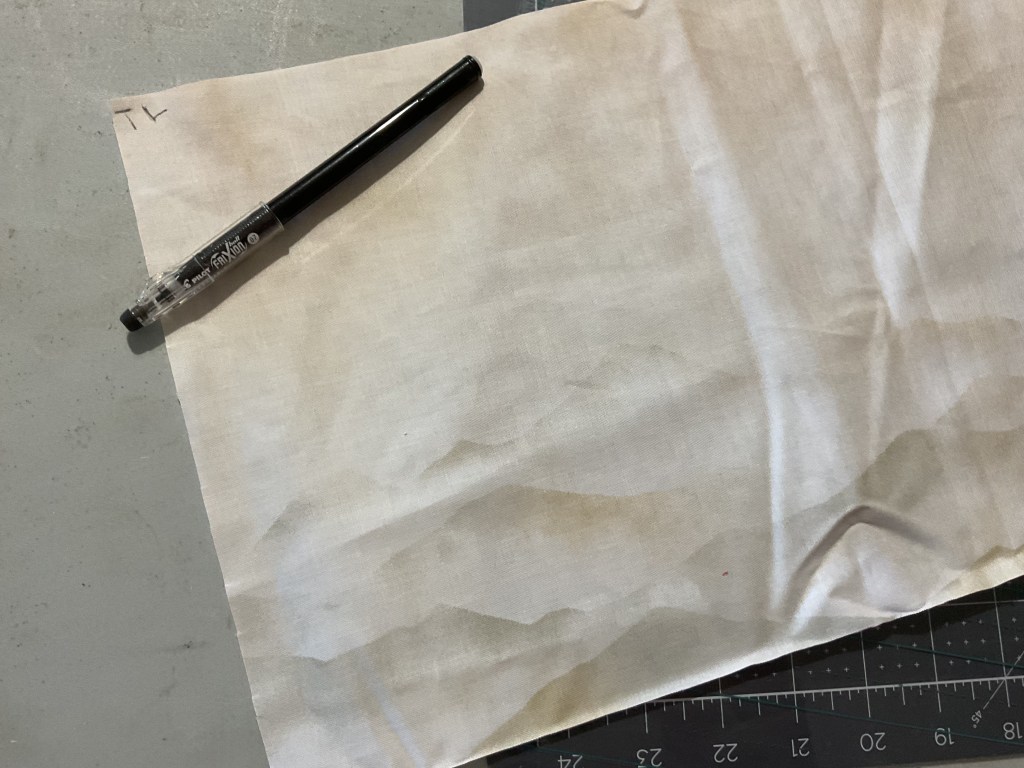A white fabric with light creases and stains, lying on a gray surface, accompanied by a black pen.