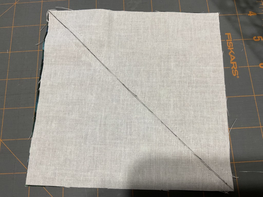 A square piece of light fabric, partially stitched diagonally, placed on a gray cutting mat with grid lines.