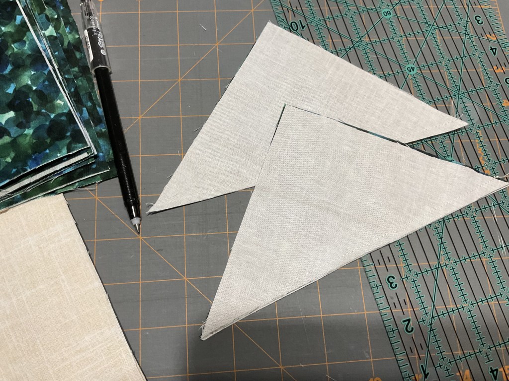 Two triangular fabric pieces cut from a beige fabric, placed on a cutting mat with grid lines, alongside a stack of green patterned fabric squares and a black pen.
