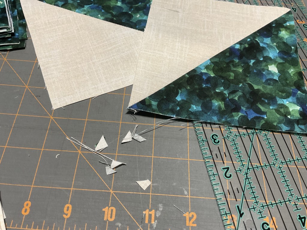 Cut fabric pieces for quilting, featuring a green and blue patterned triangle next to a light beige triangle, with small scraps of fabric and threads scattered on a cutting mat.