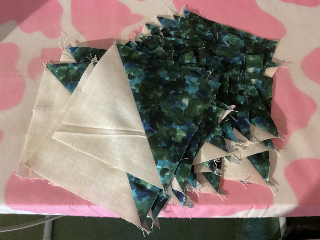 A collection of fabric pieces in various shades of green and beige, arranged in a pile on a pink and white patterned surface.